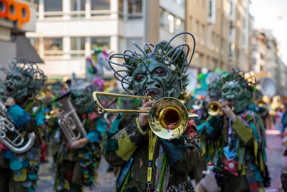 Guggenmusiker mit grüner Monster-Grende spielt Posaune, dahinter weitere Grende mit Blech am Umzug der Luzerner Fasnacht