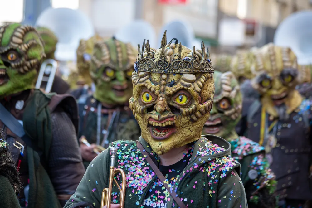 Guggenmusiker mit goldener Echsen-Grende und Krone hält die Trompete, voller Konfetti, dahinter die ganze Gruppe in Grende